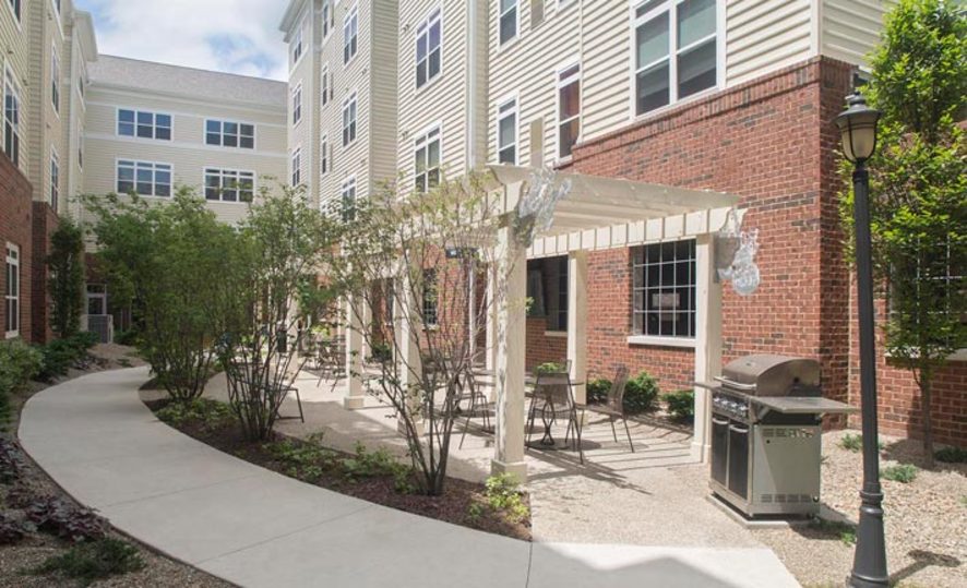sidewalk along short trees next to a seating area with pergola